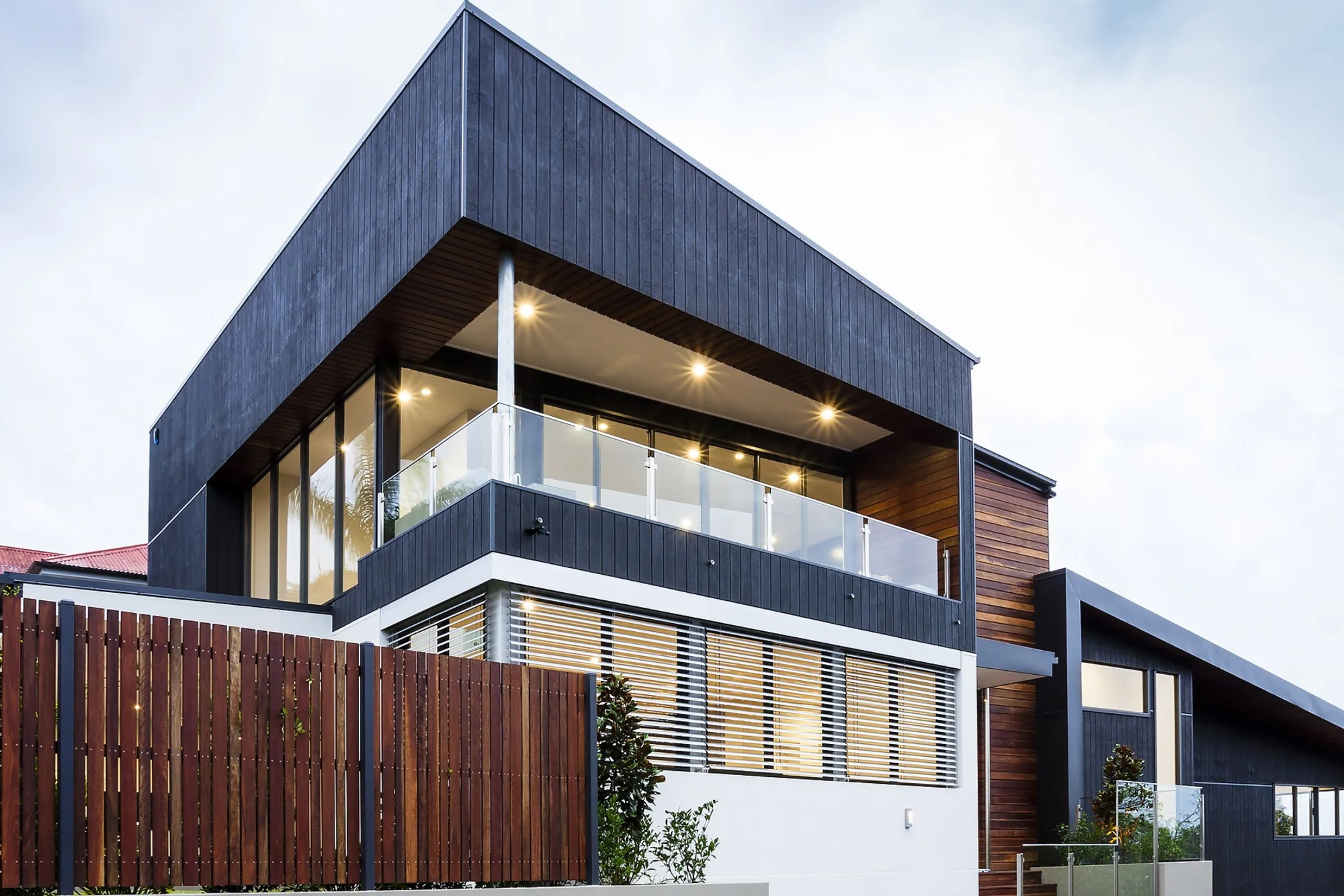 Ein sehr modernes, kubisches Einfamilienhaus mit einer großen überdachten und beleuchteten Terrasse im ersten Stock. Das Haus ist mit dunkelgrauem Holz verkleidet und von einem hohen braunen Holzzaun umgeben.