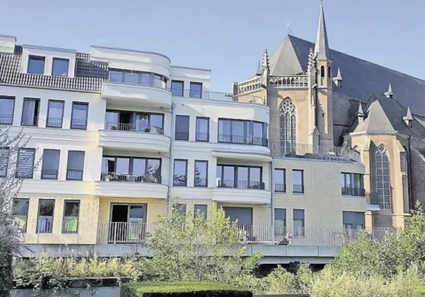 Ein großes helles Mehrfamilienhaus mit vier Etagen und vielen, hohen Glasfenstern vor einer Kirche mit spitzen Türmen.