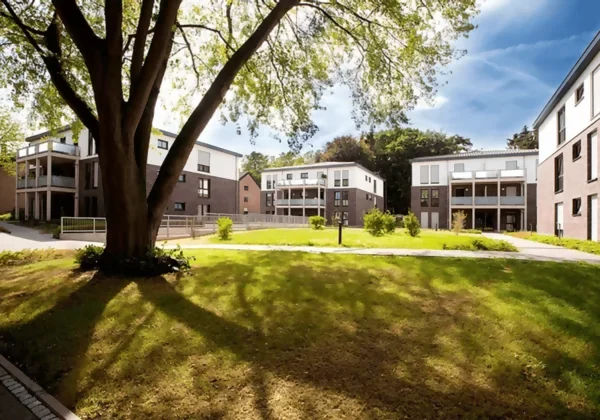 Mehrere Gebäude gruppieren sich um einen gepflegten Park, in dem ein großer grüner Baum steht.